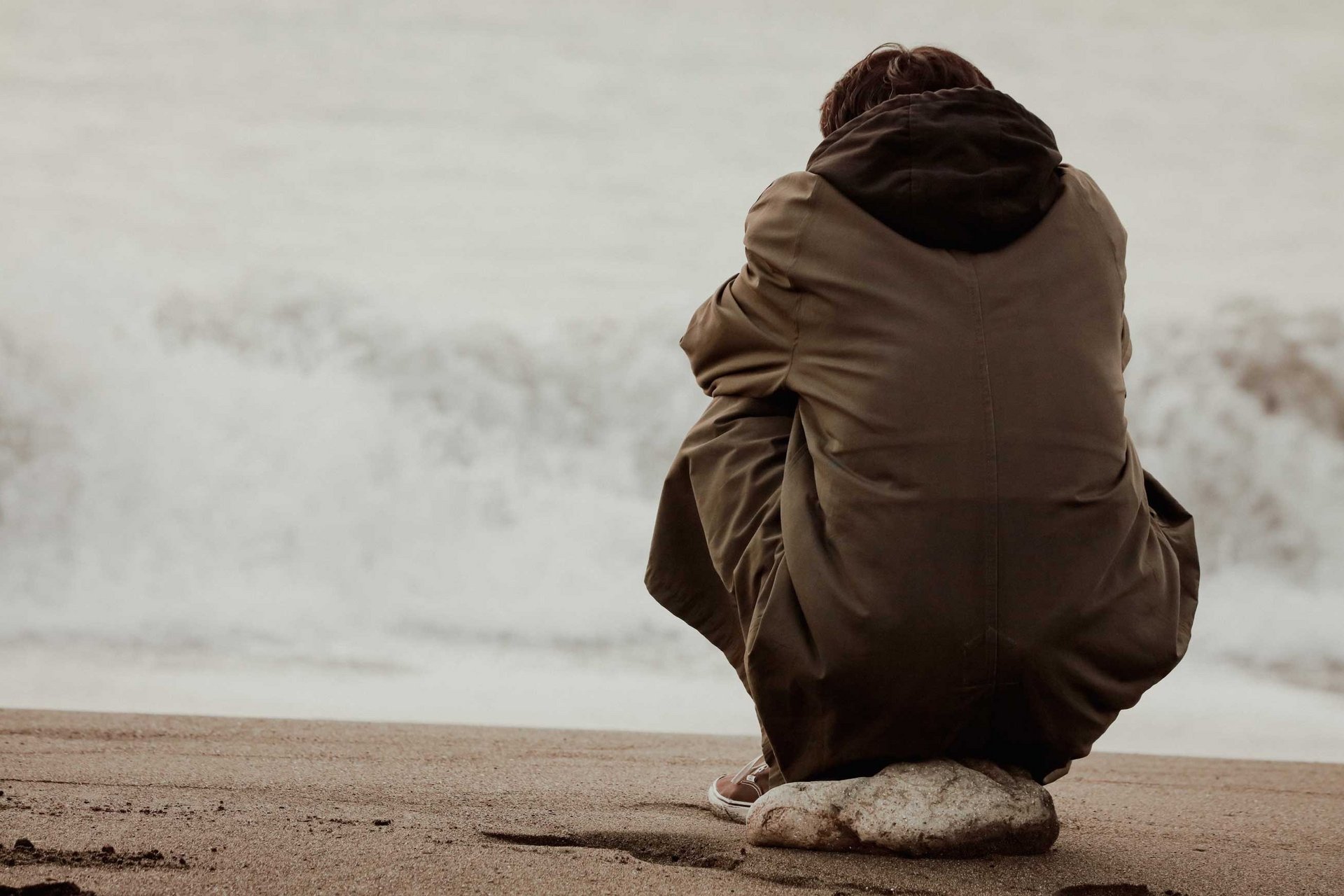 Ein Mann sitzt alleine auf einem Stein am Strand und blickt auf die Wellen hinaus.