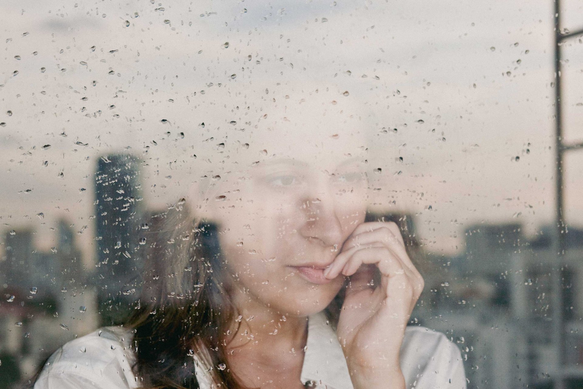 Eine Frau schaut in Gedanken verloren aus einem Fenster in den Regen und kaut an ihren Fingernägeln