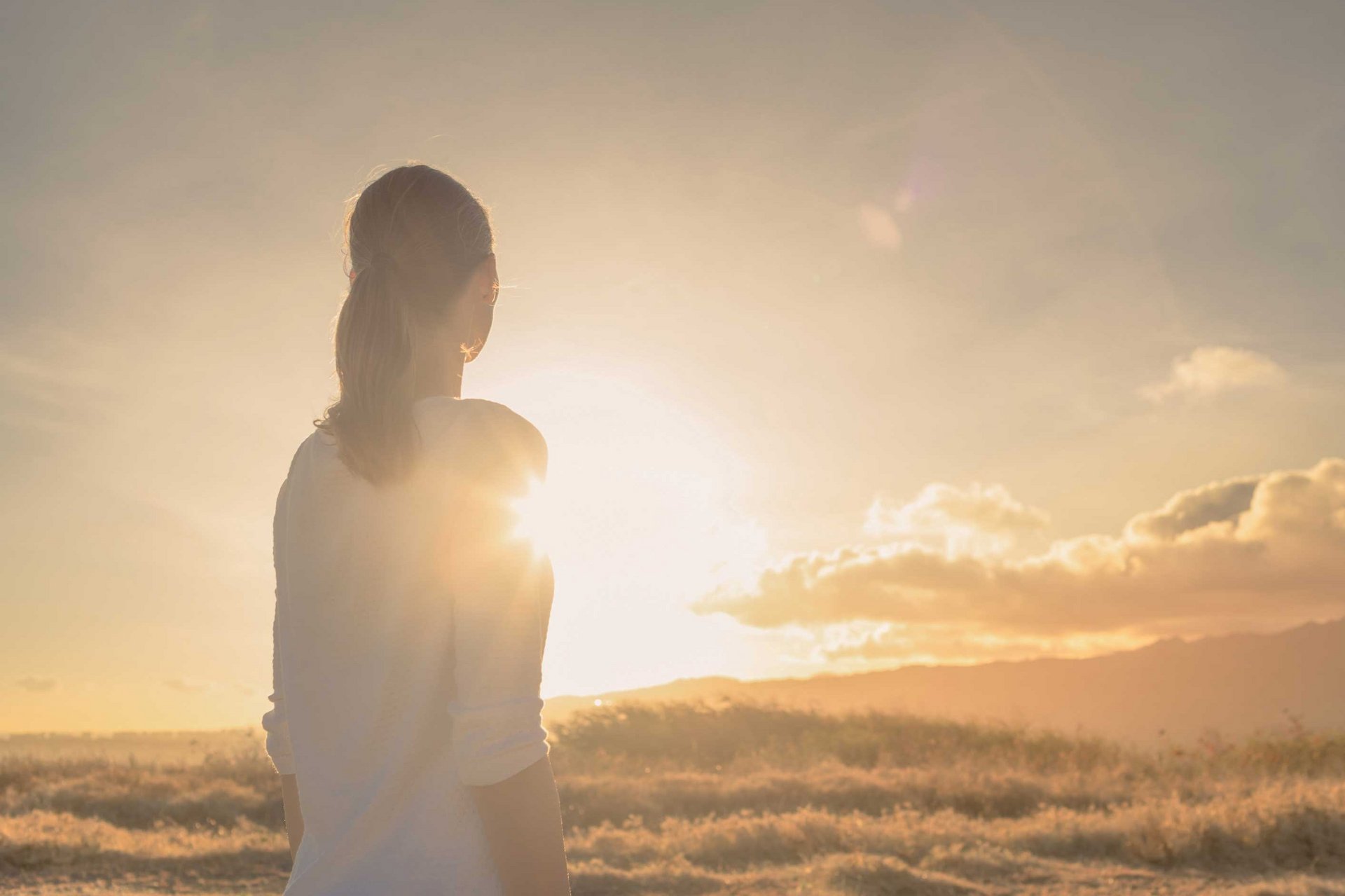 Eine Frau steht auf einem Feld und blickt in den fernen  Sonnenuntergang.