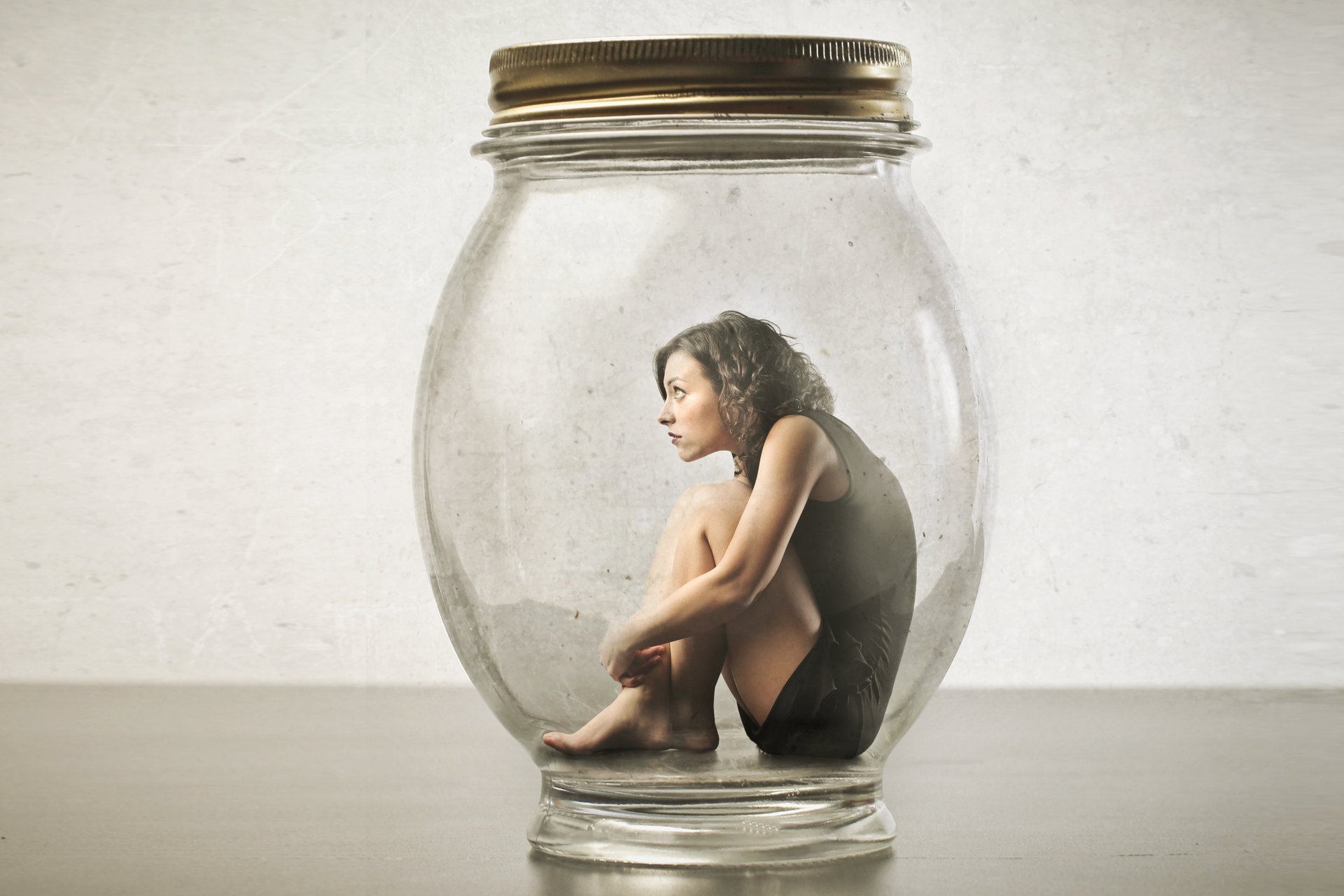 Eine Frau sitzt zusammengekauert auf dem Boden eines verschlossenes Einmachglases. 