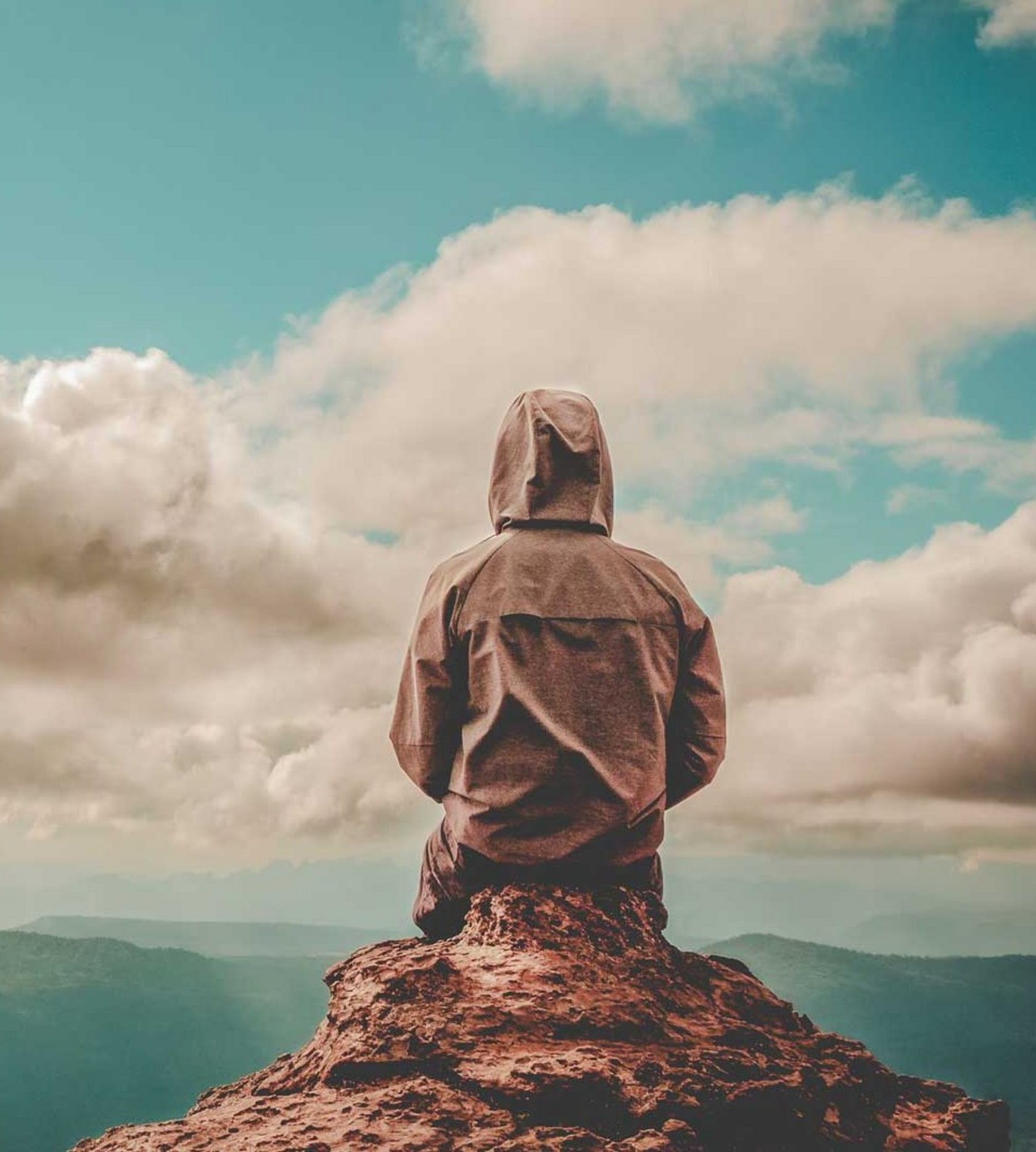 Person mit Jacke und aufgezogener Kapuze sitzt auf einem Berg und schaut in die Ferne, um Leere zu symbolsisieren