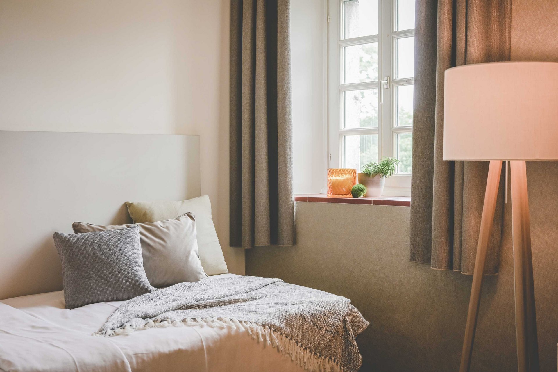 Ein Bett mit Kissen steht in einem wohnlichen Raum neben einem Fenster mit Vorhängen und einer Stehlampe.