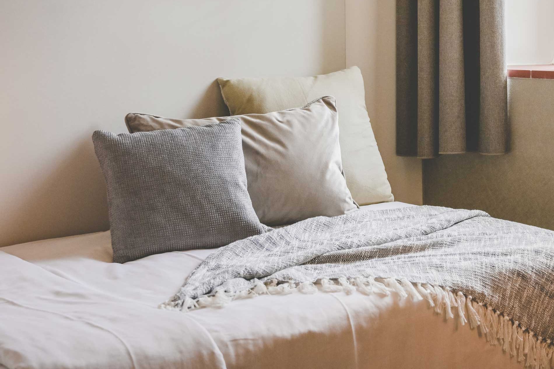 Ein Bett mit Kissen in Erdtönen steht in einem wohnlichen, lichtdurchfluteten Raum neben einem Fenster mit Vorhängen
