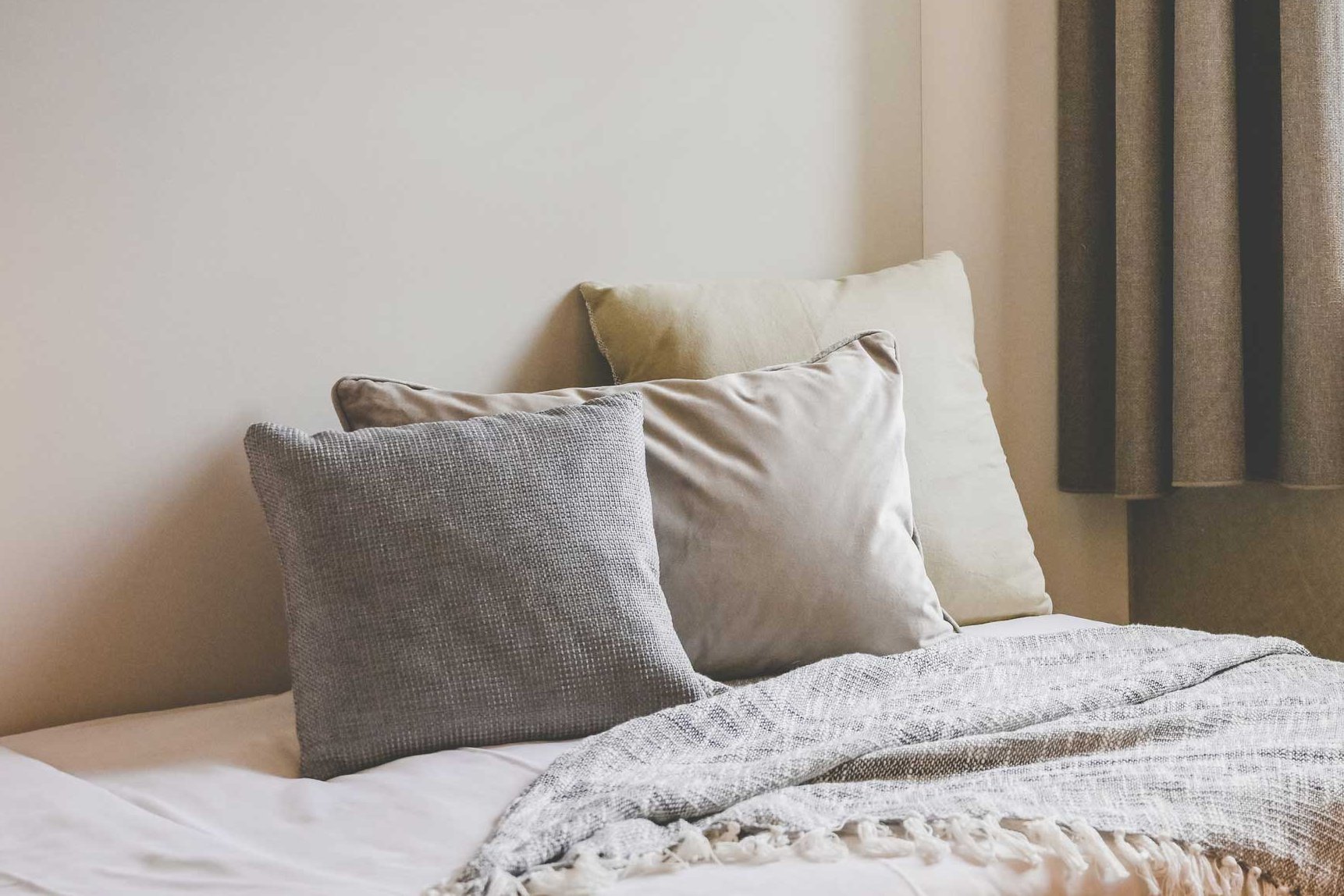 Ein Bett mit Kissen in Erdtönen steht in einem wohnlichen, lichtdurchfluteten Raum neben einem Fenster mit Vorhängen