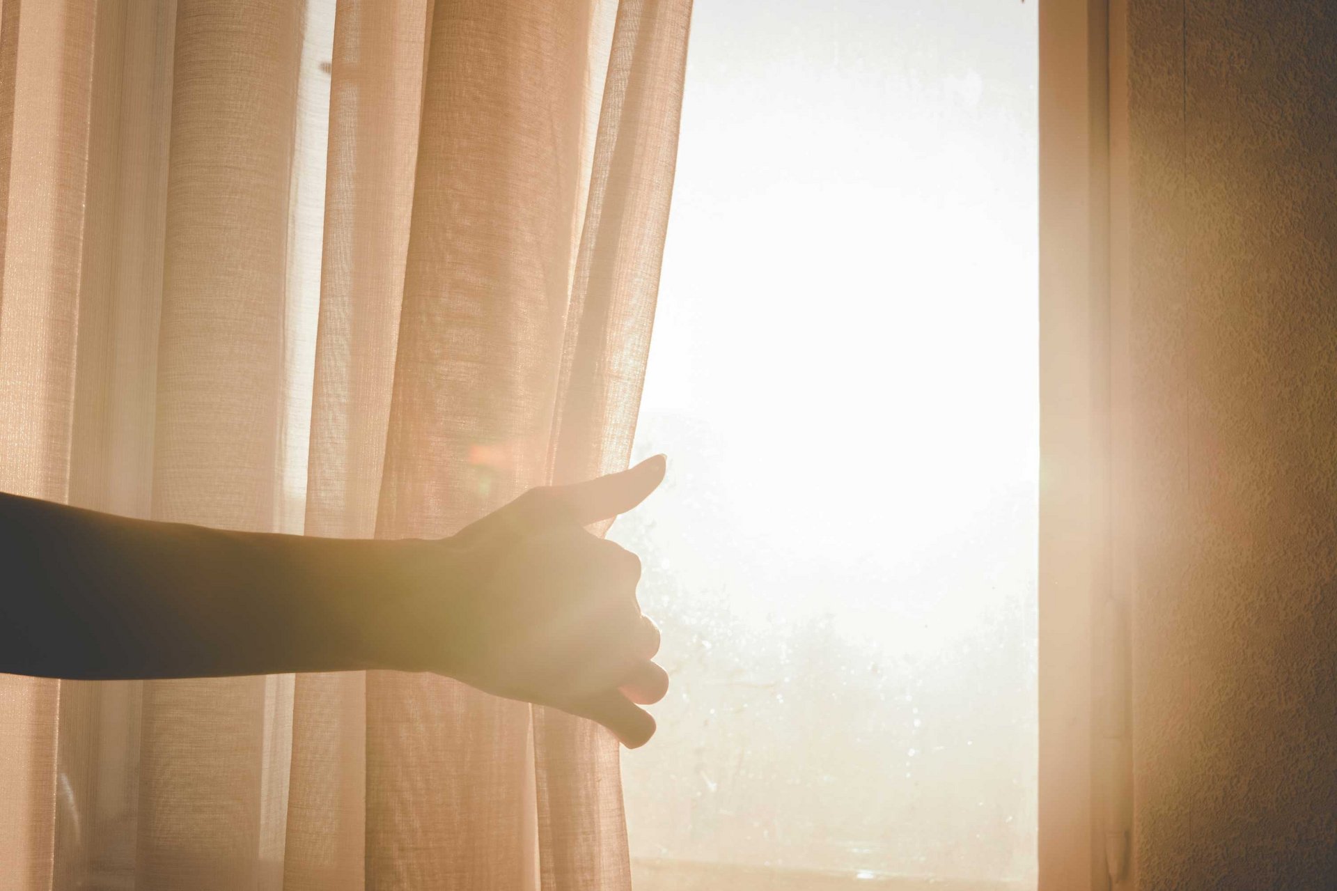 Eine Hand zieht einen Vorhang von einem von Sonnenlicht durchfluteten Fenster